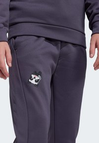 Person wearing dark purple pants and sweatshirt with a small pixel art dog graphic on the left thigh, hands relaxed by the sides.