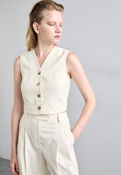 Top cropped sans manches de couleur crème avec un motif texturé et cinq boutons marron, associé à un pantalon large assorti.