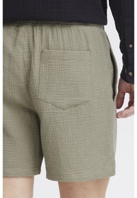 Olivgrüne Shorts aus strukturiertem Stoff mit einer sichtbaren aufgesetzten Tasche auf der Rückseite und einem elastischen Bund für Komfort.