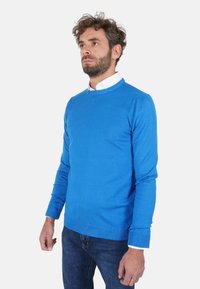Maglione azzurro lavorato a maglia con scollo rotondo, maniche lunghe e polsini e orlo a coste, indossato sopra una camicia bianca con colletto. Spessore medio.