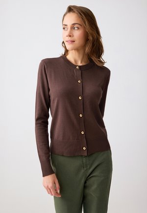 Młoda kobieta ubrana w brązowy kardigan zapinany na guziki z złotymi guzikami i zielone spodnie, patrząca na lewo na jednolitym tle.