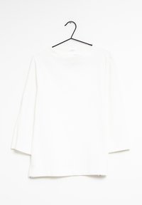 Haut blanc à manches longues avec un col rond et une coupe droite. Présente un tissu texturé et des fentes sur les côtés à l'ourlet. Suspendu à un crochet noir.