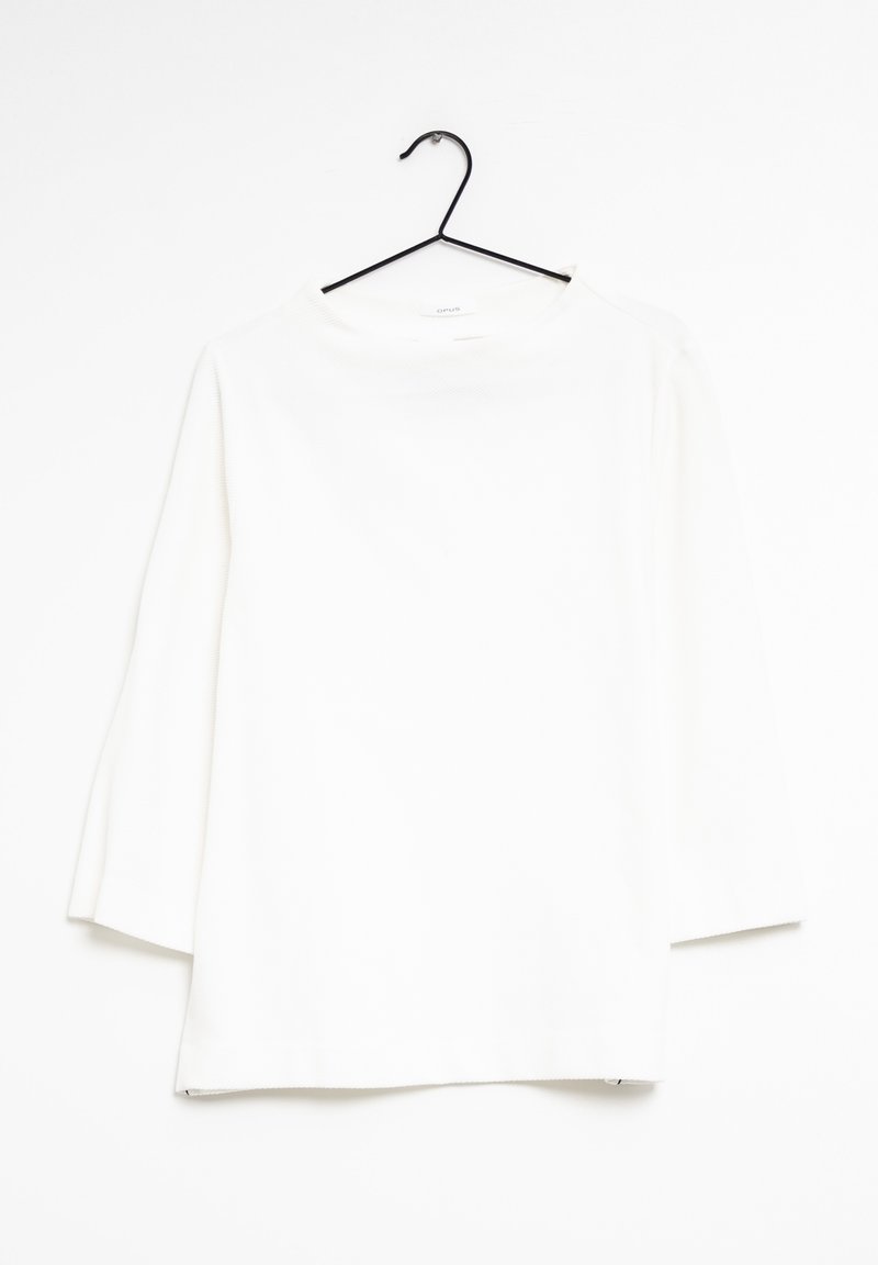 Haut blanc à manches longues avec un col rond et une coupe droite. Présente un tissu texturé et des fentes sur les côtés à l'ourlet. Suspendu à un crochet noir.