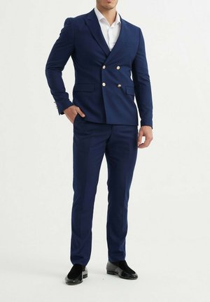 Costume bleu marine croisé avec des boutons dorés, coupe ajustée, texture lisse, associé à une chemise blanche et des chaussures noires.
