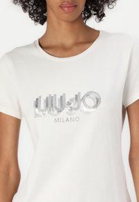 Weißes T-Shirt mit kurzen Ärmeln, verziert mit einem silbernen Strass-Logo, das "LIU JO MILANO" in einem markanten, modernen Design zeigt. Aus weichem Baumwollstoff.