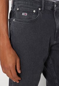 Jeans de denim negro con una textura suave, costuras visibles y un parche de logo en el bolsillo frontal. Incluye botones y remaches de metal.