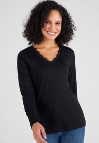 Zwarte lange mouwen top met een V-hals, voorzien van een kantafwerking. Zachte, rekbare stof met een reguliere pasvorm, gecombineerd met blauwe jeans.