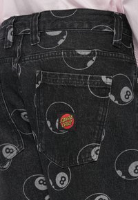 Zwarte denim jeans met een overal grijs achtbalpatroon, voorzien van een rood en geel "Santa Cruz" etiket op de achterzak.