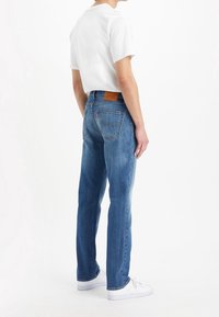 Jeans de mezclilla azul con un corte recto, que presentan un parche de marca de cuero, bolsillos traseros y un ligero desgastado. Combinados con una camiseta blanca y zapatillas.