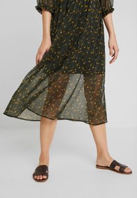 Vestido de flores de color verde oscuro, con tela transparente y un corte holgado, combinado con sandalias de tiras trenzadas de color marrón. Diseño largo y fluido.