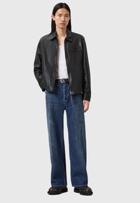Zwarte leren jas met een puntkraag, voorrits en borstzak, gedragen over een witte tanktop, gecombineerd met wijde blauwe jeans en zwarte laarzen.