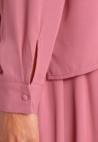 Close-up of a pink long sleeve cuff with a fabric-covered button and a small slit above the wrist on a flowing dress.