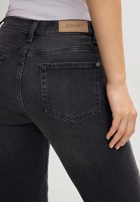 Jeans en denim noir avec un design ajusté, dotés de poches arrière, d'une étiquette en cuir de marque et de détails de couture visibles.