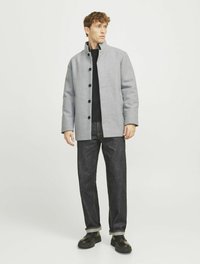 Grau meliertes Wollmantel mit Stehkragen, schwarzen Knöpfen, über einem schwarzen Hemd getragen, kombiniert mit dunkelblauen Jeans und schwarzen Turnschuhen.