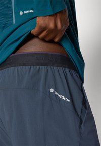 Τιρκουάζ μπλουζάκι με μακριά μανίκια και λεπτομέρειες  από πλέγμα, συνδυασμένο με γκρι σορτσάκι DuraStretch. Η μαύρη ζώνη έχει μωβ λεπτομέρειες και λογότυπο.