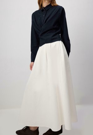 Vrouw draagt een zwarte blouse met lange mouwen en knopen, een witte zwierige rok en donkere schoenen tegen een effen lichte achtergrond.