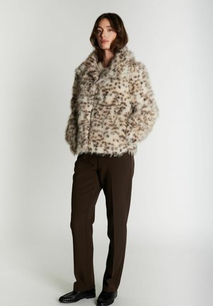 Femme portant une veste en fausse fourrure marron clair tachetée, un pantalon marron foncé et des chaussures noires, se tenant debout les mains dans les poches devant un fond uni.