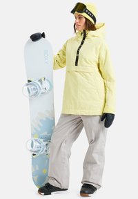 Vrouw in een gele jas en beanie staat met een witte en blauwe Roxy snowboard met bindingen, gekleed in zwarte handschoenen en sneeuwlaarzen.