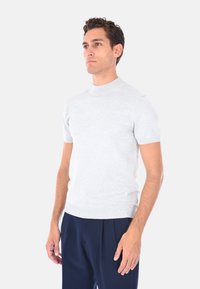 Uomo che indossa un maglione a maniche corte grigio chiaro e pantaloni scuri a pieghe blu, in piedi con le braccia rilassate su uno sfondo bianco semplice.
