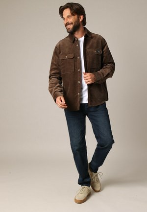 Veste en velours côtelé brun avec deux poches poitrine, fermeture à boutons et texture lisse, associée à un jean foncé et des baskets claires.