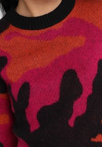 Primo piano di un maglione di lana colorato con motivi astratti rossi, rosa e neri e un colletto a coste nero.