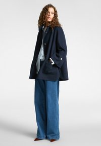 Cappotto in misto lana navy con bottoni dorati, maniche larghe e tasche anteriori, abbinato a una camicia azzurro chiaro e jeans in denim larghi.