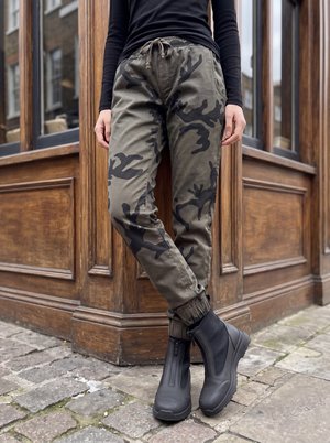 Personne portant un pantalon camouflage vert et des bottes noires debout dans une rue pavée près des coins d’un bâtiment en bois.