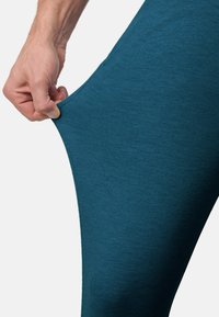 Leggings teal en tissu extensible, avec une texture lisse. Une main saisit la taille, démontrant l'élasticité et l'ajustement.