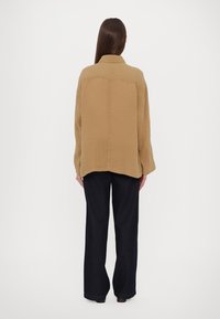 Brown collared jacket in a textured fabric, featuring a straight cut and visible stitching, paired with dark wide-leg pants.