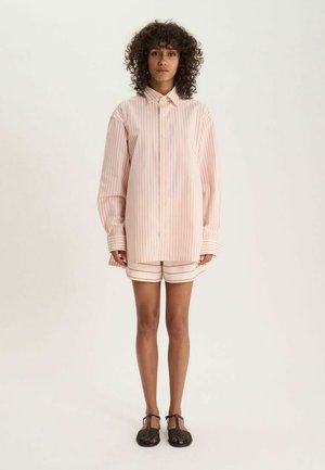 Femme aux cheveux bouclés debout face à nous, portant une longue chemise rayée rose pâle, un short assorti et des sandales tissées foncées.