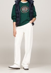 Grüner Pullover mit rundem, besticktem Logo. Dunkelblaue Steppweste über weißen, weit geschnittenen Hosen und weißen Schuhen mit schwarzen Sohlen.