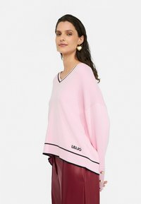 Hellrosa Sweater mit V-Ausschnitt und schwarzer Borte, ausgestattet mit einem gestickten Logo am Saum und übergroßen Ärmeln mit Knopfakzenten.