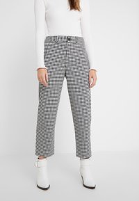 Donna che indossa pantaloni dritti a quadri bianco e nero, top a maniche lunghe bianco a coste e stivaletti bianchi con cinturini.