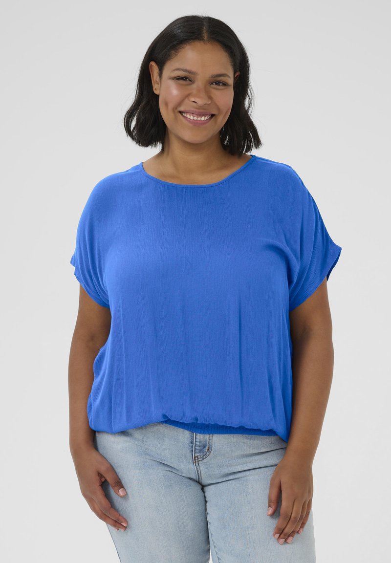 Blue short-sleeve blouse in lightweight fabric with a relaxed fit. Features a round neckline and gathers at the hem. Worn with light jeans.