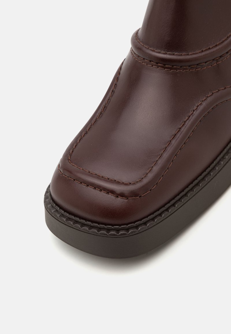 Brune læder ankelstøvle med en glat overflade, rund tå, synlige syninger og en kontrasterende mørk sål med en struktureret kant.