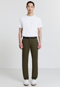 Uomo che indossa una maglietta bianca semplice, pantaloni verde oliva con una cintura nera e sneakers bianche, in piedi contro uno sfondo grigio chiaro.