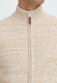 Nærbilde av en beige strikkegenser med ribbestrikket høy krage og en hvit glidelås som går ned langs midten foran.