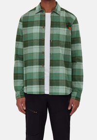 Zielona koszula flanelowa w kratę z guzikiem z przodu, z klapami i małym detalem z logo. Nosi się ją na białym T-shircie.