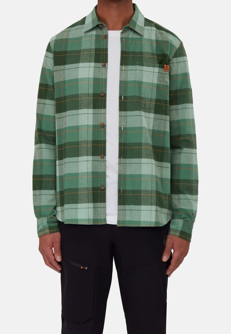 Zielona koszula flanelowa w kratę z guzikiem z przodu, z klapami i małym detalem z logo. Nosi się ją na białym T-shircie.