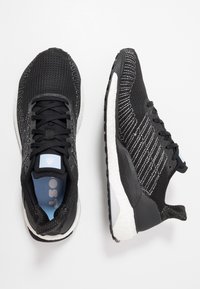 Baskets de sport noires avec un dessus en maille texturée, semelle blanche rembourrée et bandes réfléchissantes argentées sur les côtés. Semelle intérieure bleue visible.