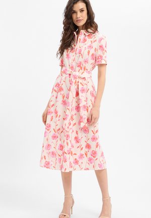 Frau trägt ein knielanges Kleid mit pinken und orangefarbenen Blumen, kurzen Ärmeln und einem Taillengürtel, kombiniert mit beigen Pumps mit Knöchelriemen.