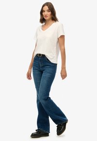 Lys beige t-shirt med v-udskæring kombineret med mørkeblå udvidede jeans. Outfitet er suppleret med et sort bælte og sorte sko.