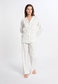 Witte pyjama set met kleurrijke patronen van bomen en versieringen. Lange mouwen met knopen en losse broekspijpen. Zachte stof.