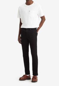 Camiseta blanca de algodón con un bolsillo en el pecho, combinada con pantalones vaqueros negros y zapatos de cuero marrón. Diseño simple y clásico con líneas limpias.