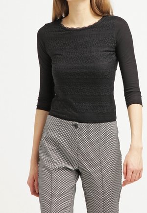 Femme vêtue d'un haut en dentelle noire à manches longues et d'un pantalon taille haute à motifs noirs et blancs, debout sur un fond clair uni.