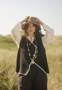Gilet noir en laine avec des finitions blanches, superposé sur une blouse blanche brodée, accompagné d'un collier de perles jaunes et d'un chapeau à large bord beige.