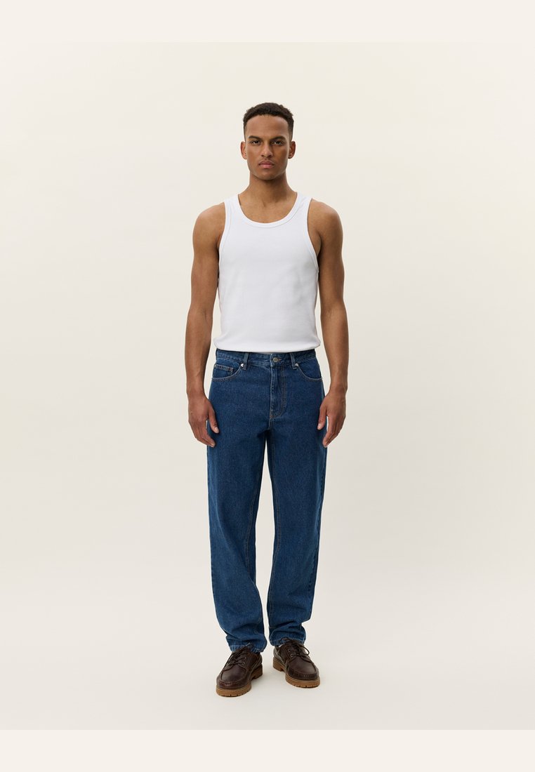 Weißes Tanktop, blaue hochtaillierte Jeans, braune Schuhe. Modell steht gerade vor einem neutralen Hintergrund und präsentiert die Kleidung und Passform.