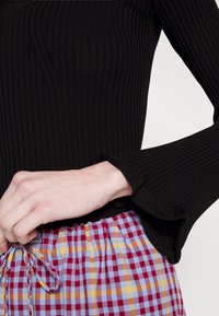 Schwarzes, geripptes Langarmshirt mit ausgestellten Bündchen, kombiniert mit einem bunten, karierten Rock mit elastischem Bund, der blaue, gelbe und kastanienbraune Quadrate zeigt.