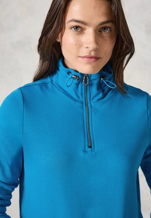 Sudadera azul con cremallera y collar alto, textura suave y detalles con cordón. Presenta una cremallera de cuarto de longitud en la parte frontal.