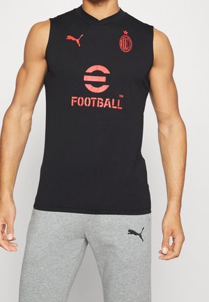 Haut de football sans manches noir de l'AC Milan avec logo rouge Puma et marquage eFootball porté par une personne debout en pantalon de survêtement gris Puma.
